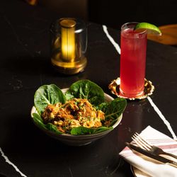 A bowl of crispy fried chicken pieces on baby spinach leaves paired with a tall pink cocktail garnished with a lime wedge.