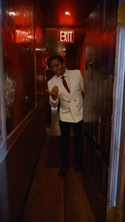 A man in a white double-breasted blazer and red tie stands in a narrow hallway under a red EXIT sign, inside Room 207.