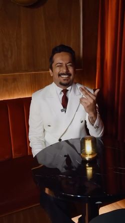 Man in a white double-breasted jacket and red tie sitting at a cozy bar booth with red upholstery and a small candle on a gle