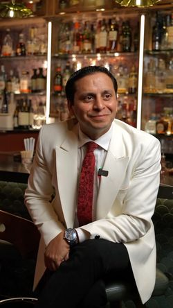 Man in a white blazer and red tie seated in a cozy bar with shelves of classic and vintage cocktails behind him