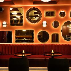 Cozy bar interior at Room 207 with red velvet seating, round tables, and decorative varied mirrors on warm orange walls.