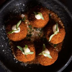 Five breaded and fried appetizer balls topped with a dollop of creamy white sauce and small fresh green herbs on a dark plate