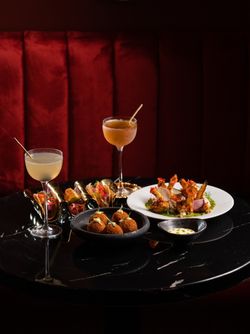 Cocktail bar setup with classic cocktails and appetizer plates on a black table against a red booth backdrop.