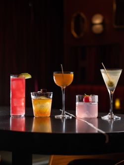 Five classic and craft cocktails arranged on a black reflective bar table in a cozy, dimly lit speakeasy setting.