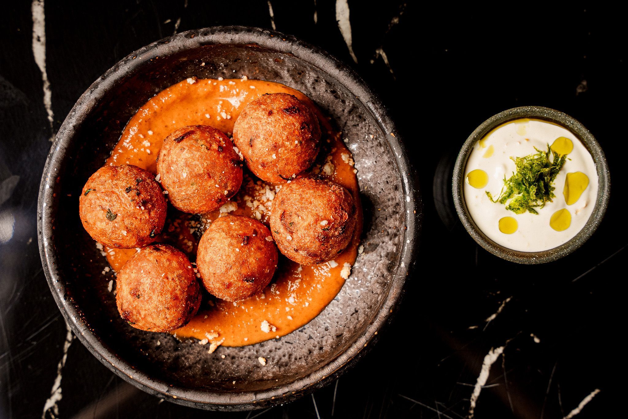 Seven round fried appetizers served on a black plate with orange sauce, accompanied by a small bowl of white dipping sauce.