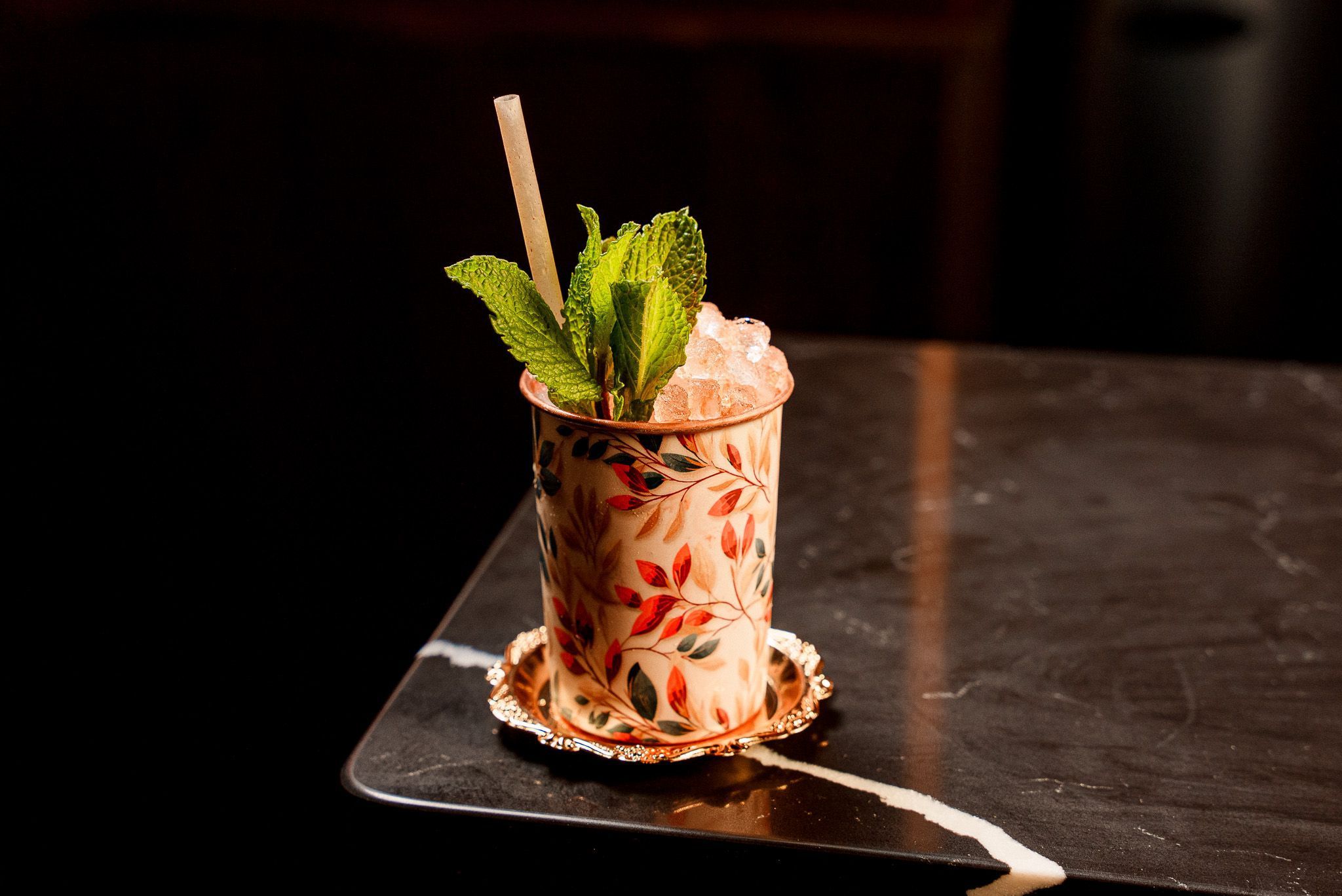 A craft cocktail garnished with fresh mint served in a decorative floral cup on a dark marble table.